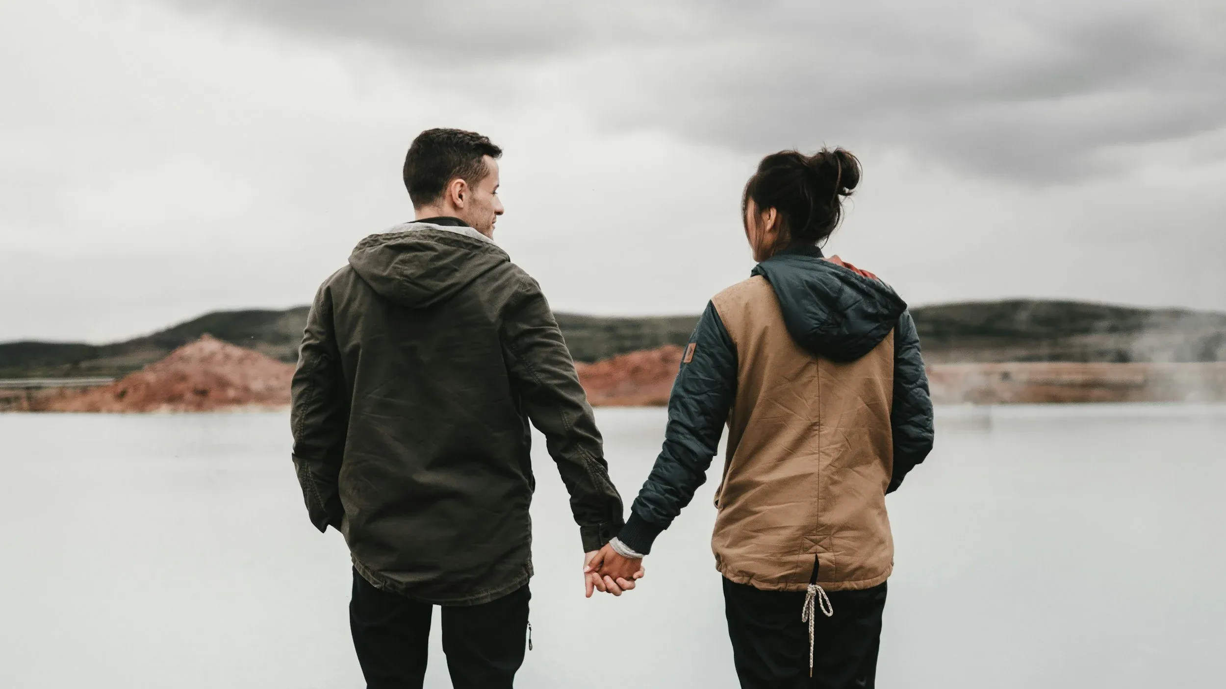 A couple seen from behind, holding hands and looking out over a lake under a cloudy sky
