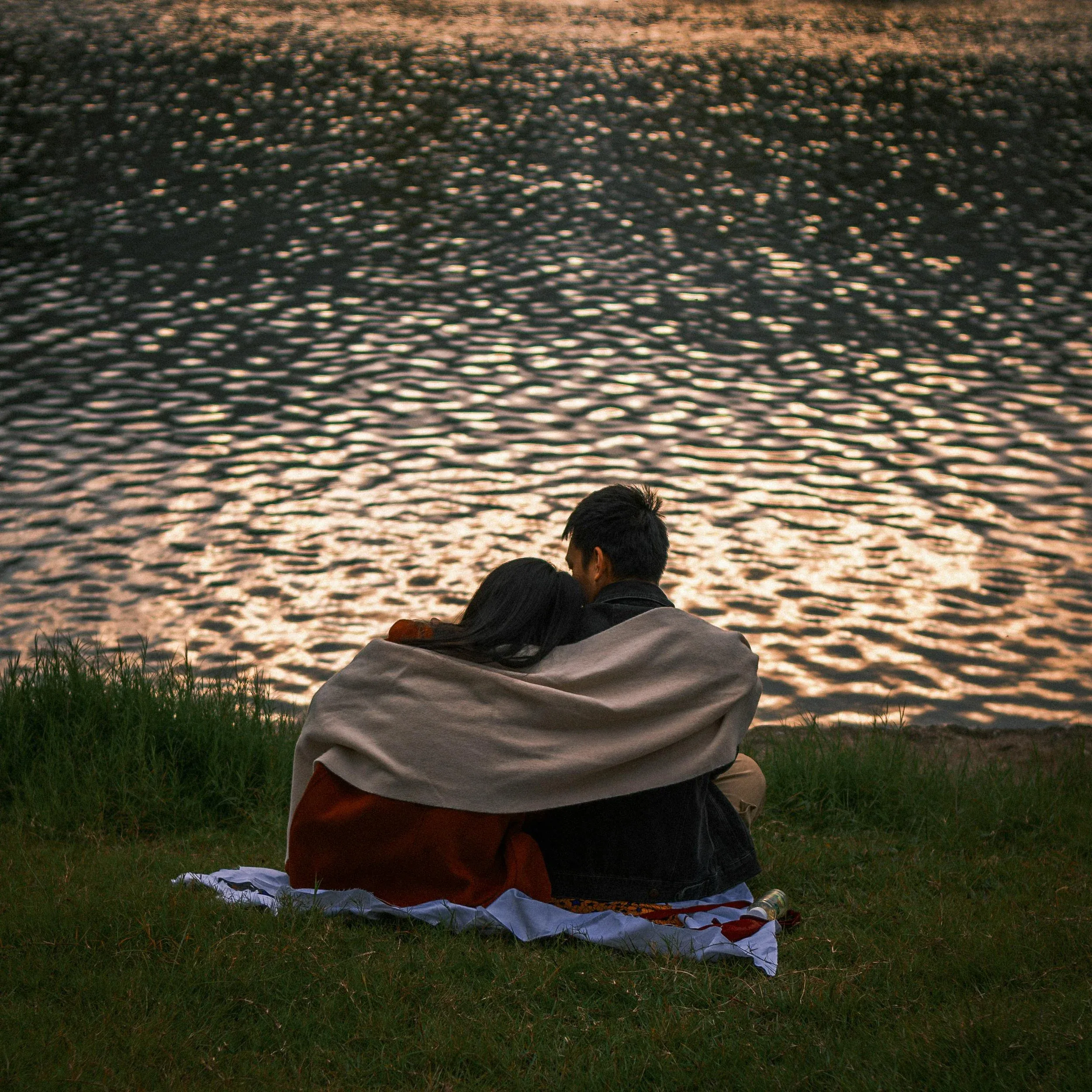 A couple wrapped in a blanket, sitting on the grass by the water at dusk