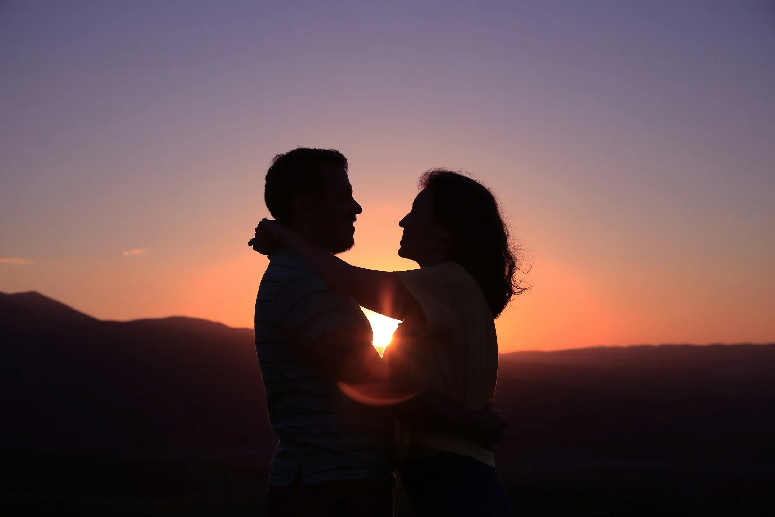 A couple embracing, silhouetted against a purple and orange sunset over hills