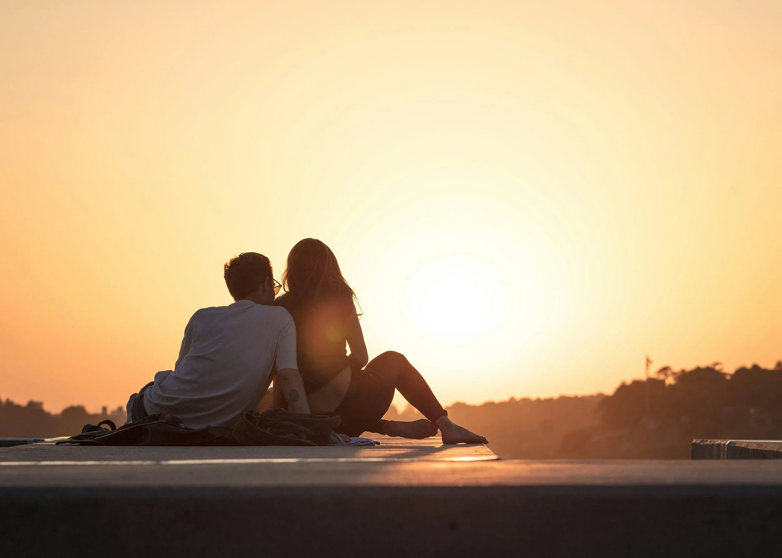 A couple sitting side by side on a ledge, silhouetted against a sunset